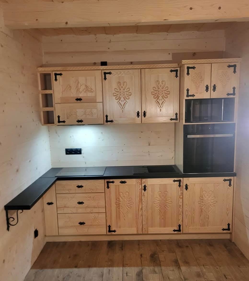 Drewniana kuchnia z rzeźbionymi frontami i czarnym blatem, stylizowana na góralski, z widoczną płytą grzewczą i piekarnikiem w zabudowie.