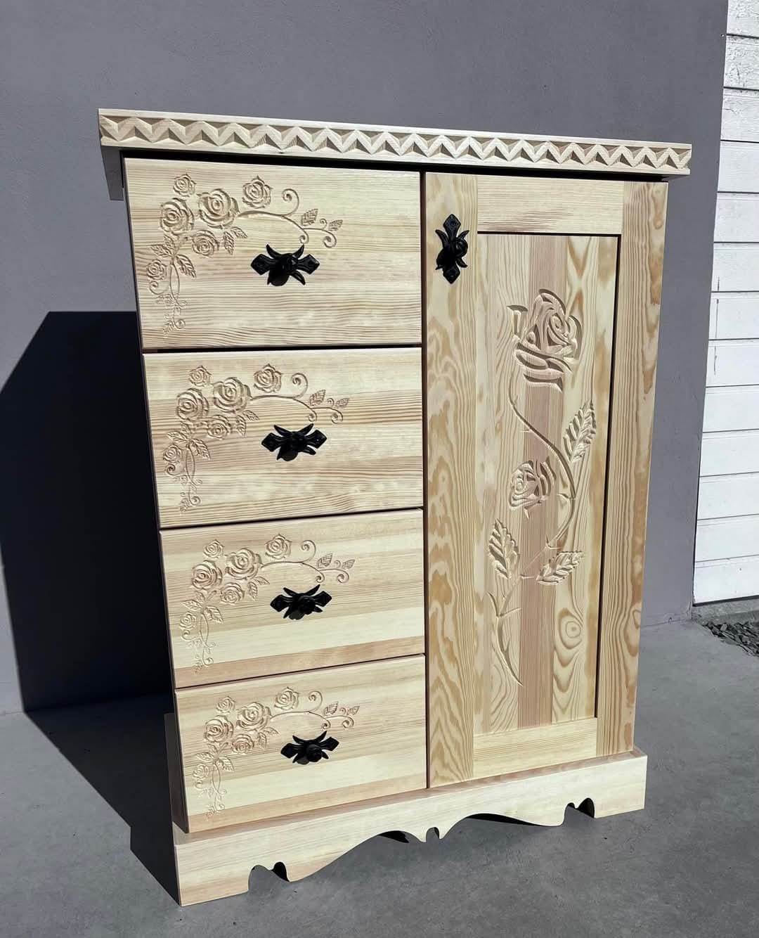 Drewniana szafa z szufladami, zdobiona motywem róż i kwiatów, z ciemnymi uchwytami. Widoczne detale snycerskie i unikalne usłojenie drewna.