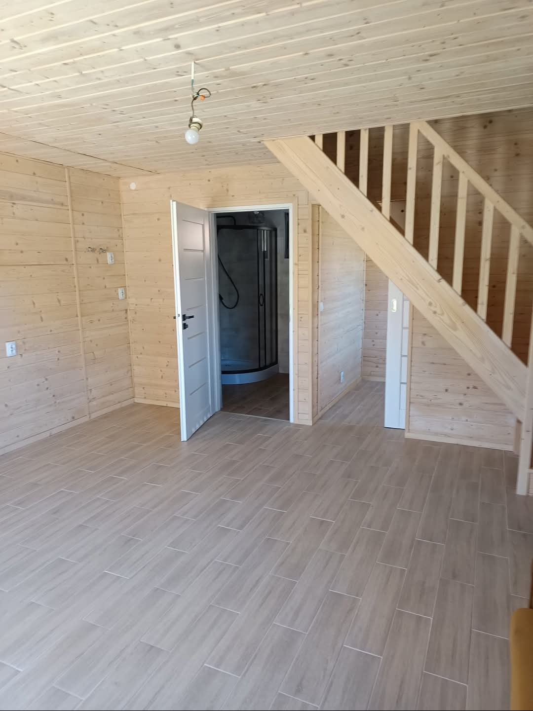Jasne wnętrze z drewnianymi ścianami, schodami i podłogą z płytek. Otwarta łazienka z kabiną prysznicową. Widoczny surowy kabel z żarówką.