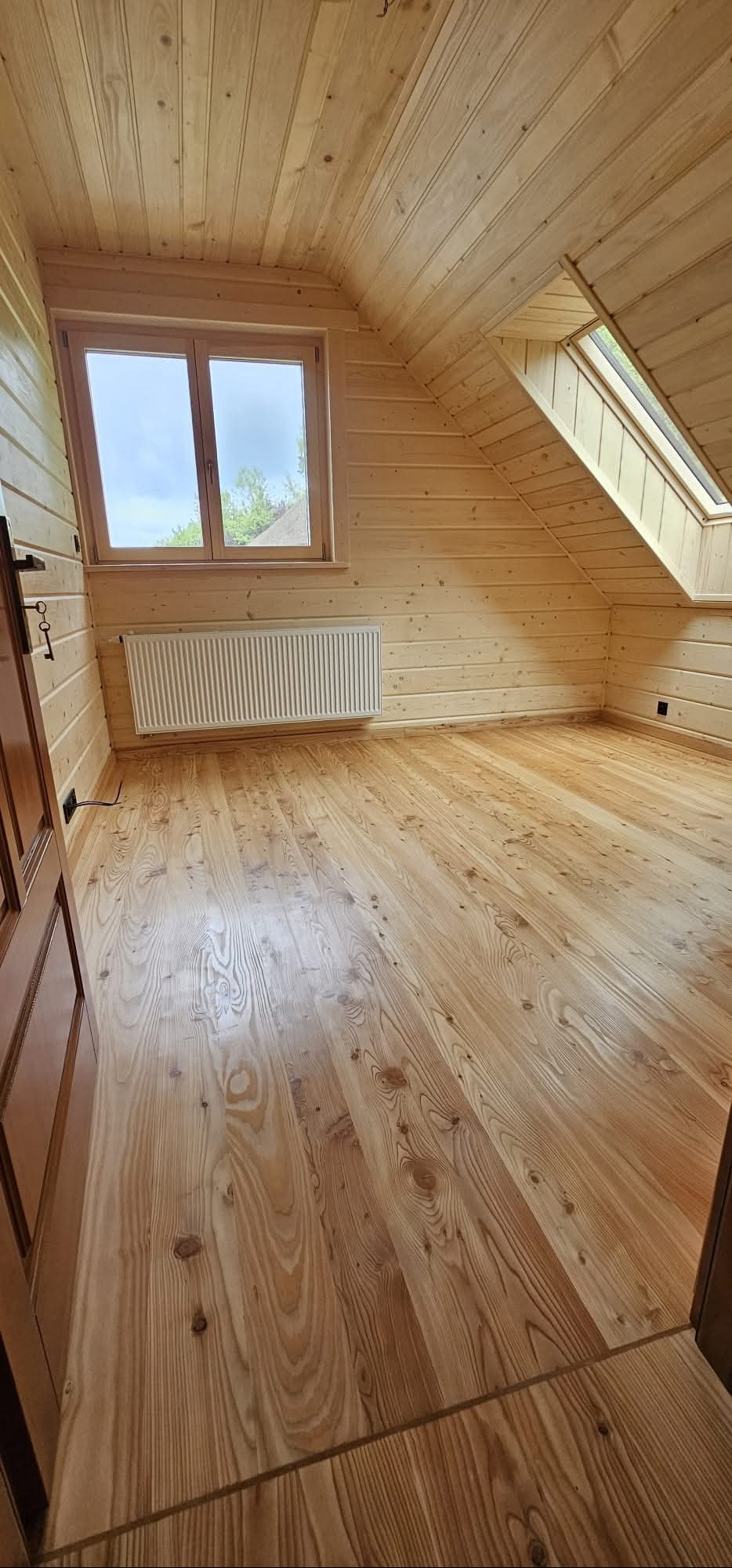 Jasne drewniane wnętrze poddasza z oknami: standardowym i dachowym. Widoczne deski na ścianach, podłodze i suficie, grzejnik pod oknem. Naturalne wykończenie.