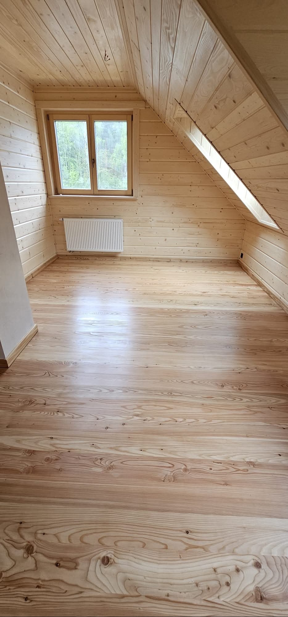 Jasne, drewniane wnętrze poddasza ze skośnym sufitem, oknem i grzejnikiem. Świeżo wykończona podłoga z wyraźnym rysunkiem słojów, ciepłe oświetlenie.
