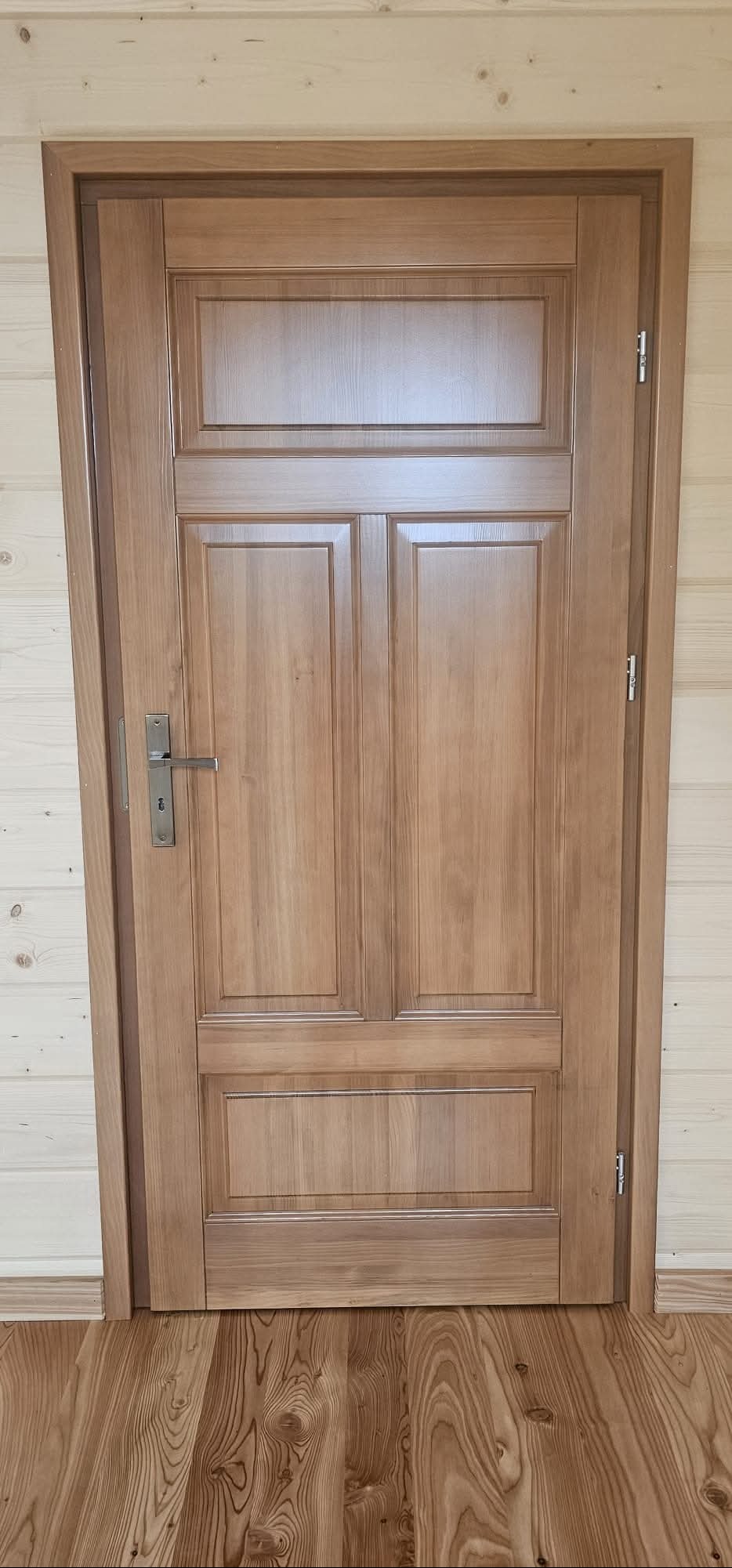 Drewniane, lakierowane drzwi wewnętrzne z ozdobnymi panelami, zamontowane w pomieszczeniu wykończonym boazerią i drewnianą podłogą. Widoczny zamek i zawiasy.