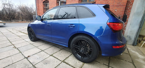 Niebieski SUV Audi Q5 z przyciemnionymi szybami i czarnymi felgami stoi na kostce brukowej przed ceglanym budynkiem w pochmurny dzień.