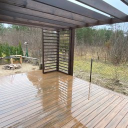 Konstrukcje Drewniane Błażej Gregus - Zadaszony taras z deski kompozytowej w kolorze ciemnego drewna, z widokiem na ogród wczesną wiosną, z elementami pergoli i paleniskiem w tle.