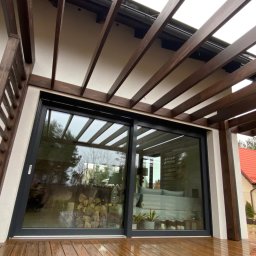 Konstrukcje Drewniane Błażej Gregus - Zadaszony taras z drewnianą pergolą i schodami, widok na przesuwne drzwi tarasowe i ogród z domem w tle.