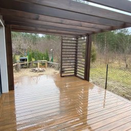 Konstrukcje Drewniane Błażej Gregus - Zadaszony taras z drewnianą pergolą i ażurową ścianką, po deszczu, z widokiem na ogród z grillem i stertą drewna.