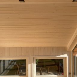 Konstrukcje Drewniane Błażej Gregus - Wnętrze jasnej altany z drewnianą boazerią, widoczne okna z odbiciem otoczenia i punktowe oświetlenie sufitowe.