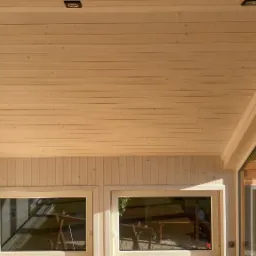 Wnętrze jasnej altany z drewnianą boazerią, widoczne okna z odbiciem otoczenia i punktowe oświetlenie sufitowe.