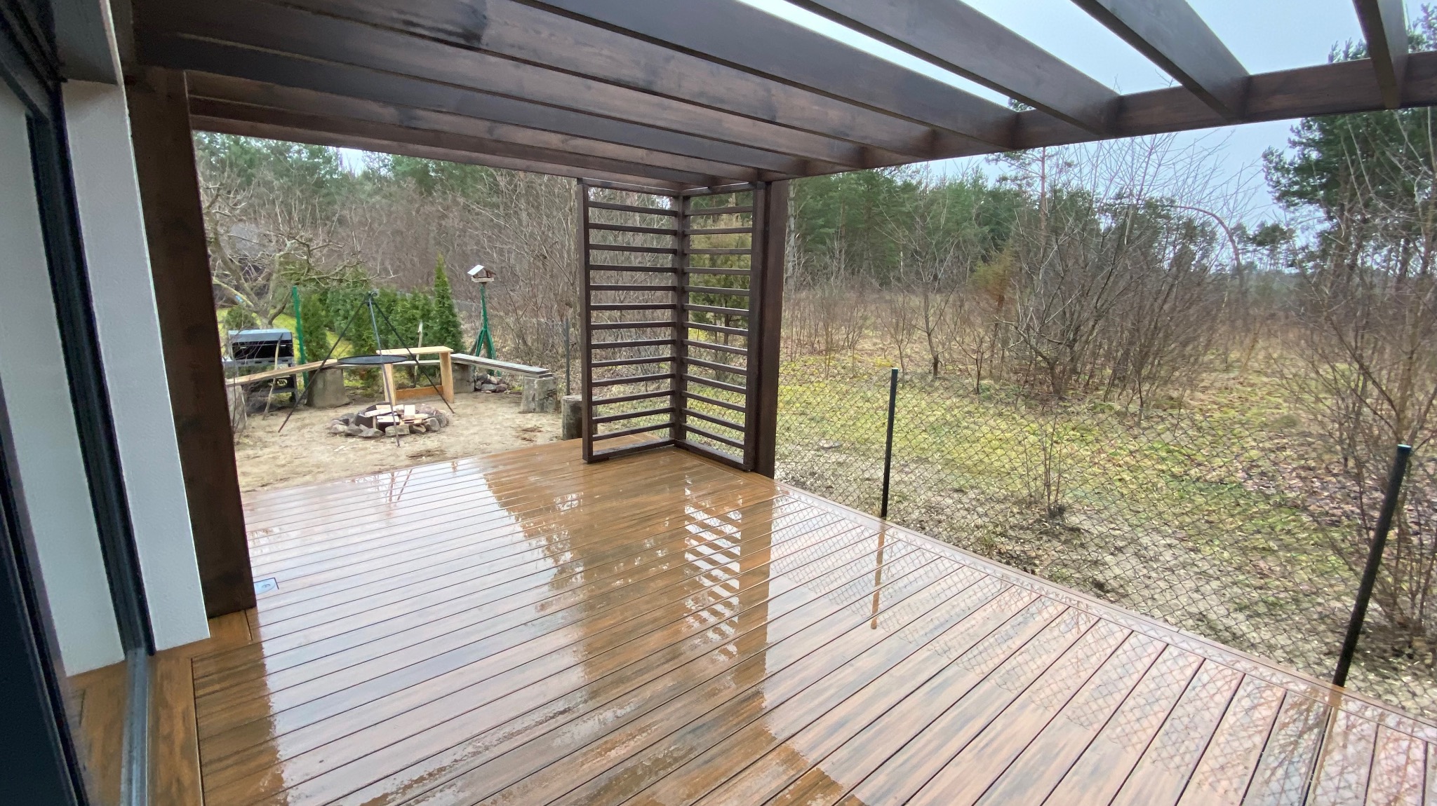 Zadaszony taras z deski kompozytowej w kolorze ciemnego drewna, z widokiem na ogród wczesną wiosną, z elementami pergoli i paleniskiem w tle.