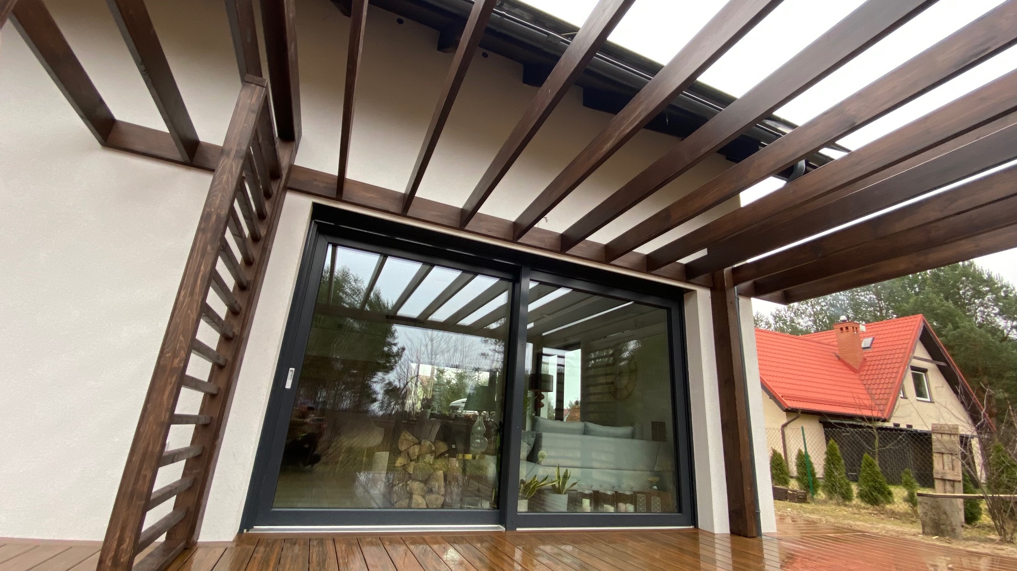 Zadaszony taras z drewnianą pergolą i schodami, widok na przesuwne drzwi tarasowe i ogród z domem w tle.