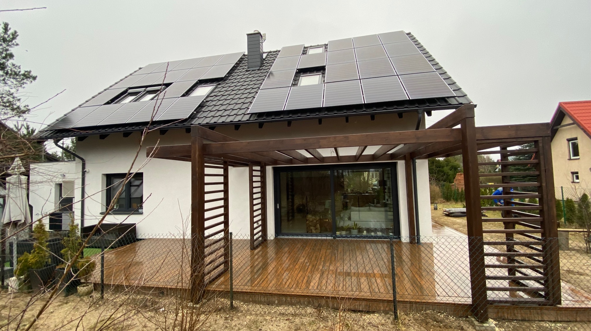 Nowoczesny dom z białymi ścianami, panelami słonecznymi na dachu i drewnianą pergolą z tarasem, widok z zewnątrz w pochmurny dzień.