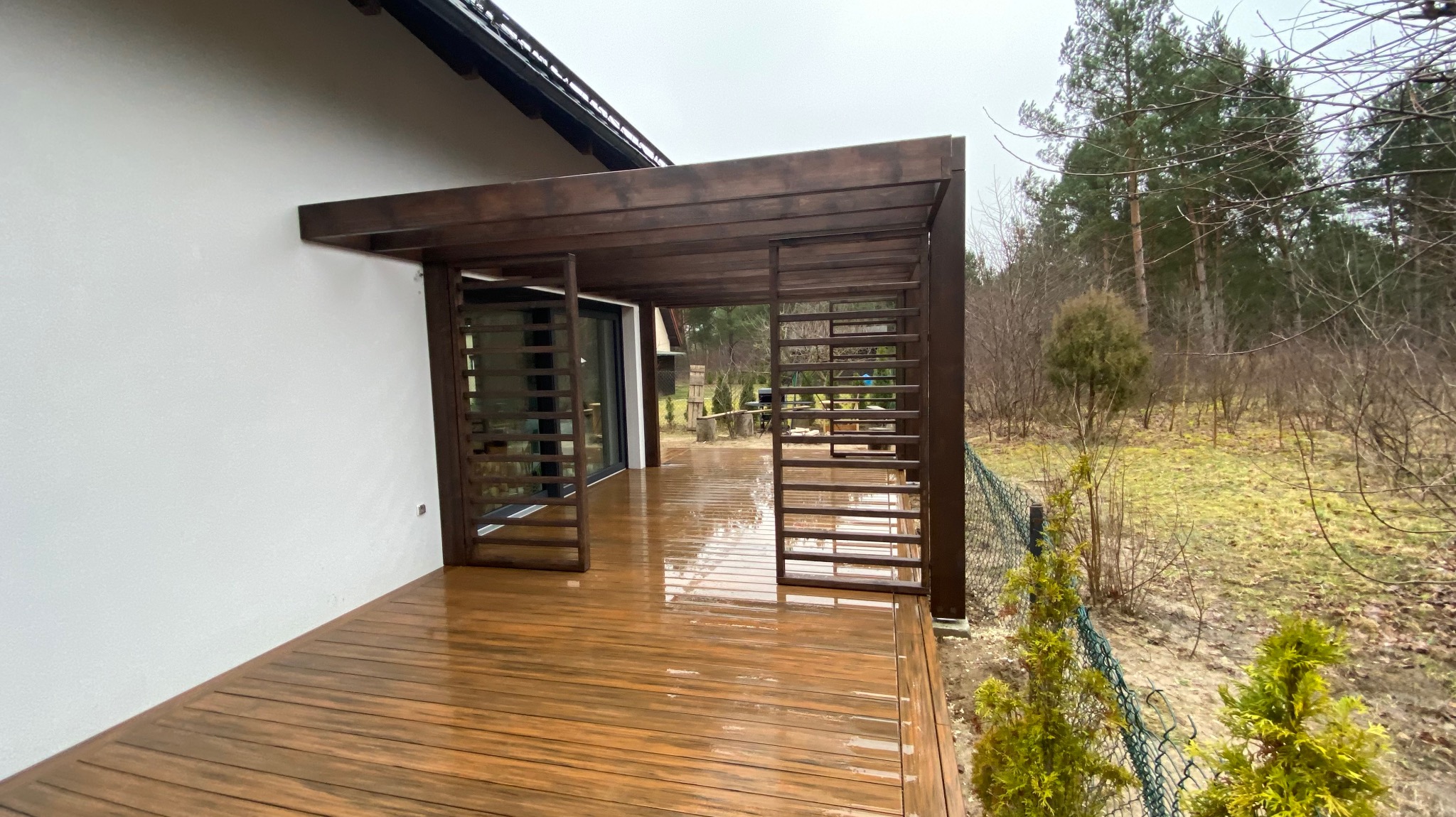 Zadaszony taras z drewnianą pergolą i podłogą z desek kompozytowych, po deszczu, widok na ogród z młodymi drzewkami iglastymi.