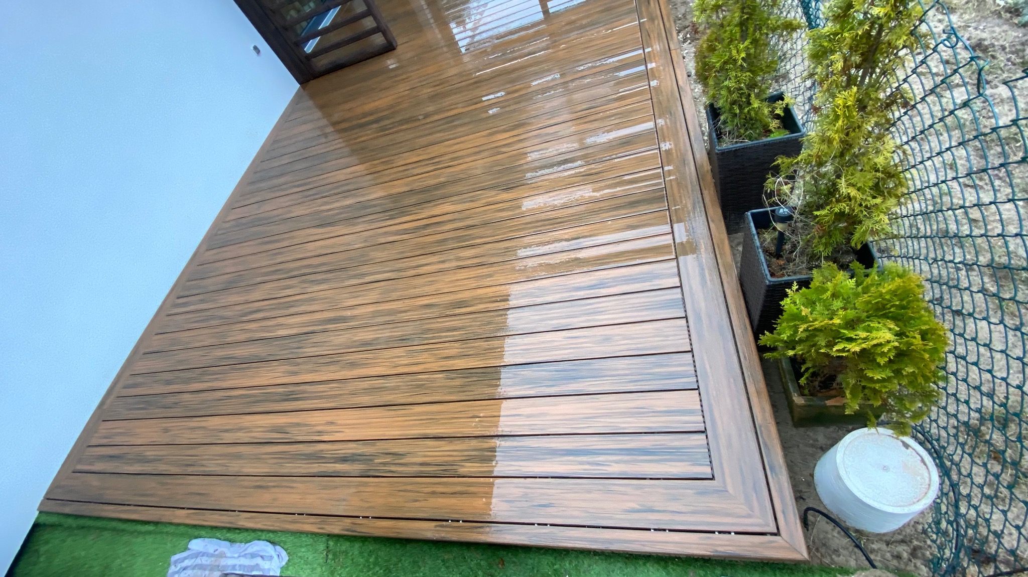Świeżo umyty taras z desek kompozytowych w odcieniach brązu, obok donice z iglakami, widoczna siatka ogrodzeniowa i fragment białej ściany budynku.