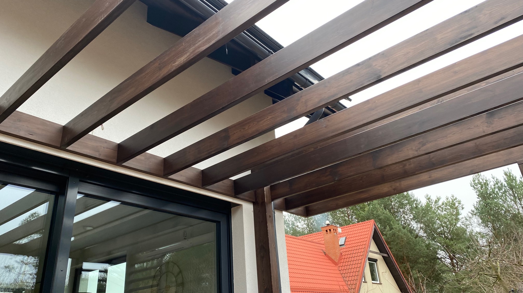Ciemnobrązowa, drewniana pergola przylegająca do białego budynku z dużym oknem przesuwnym, widok z dołu na konstrukcję i fragment dachu sąsiedniego domu z czerwoną dachówką w tle.