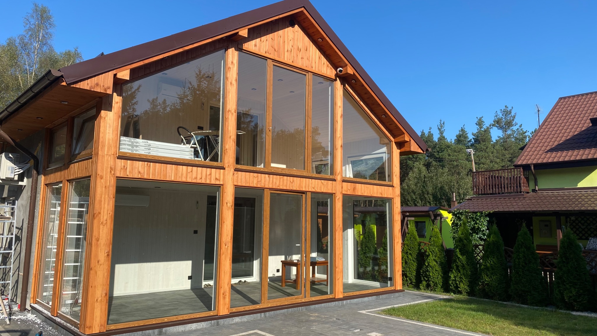 Nowoczesny dom z drewna z dużymi przeszkleniami, widoczny fragment elewacji i otoczenie, na trawniku rosną tuje.