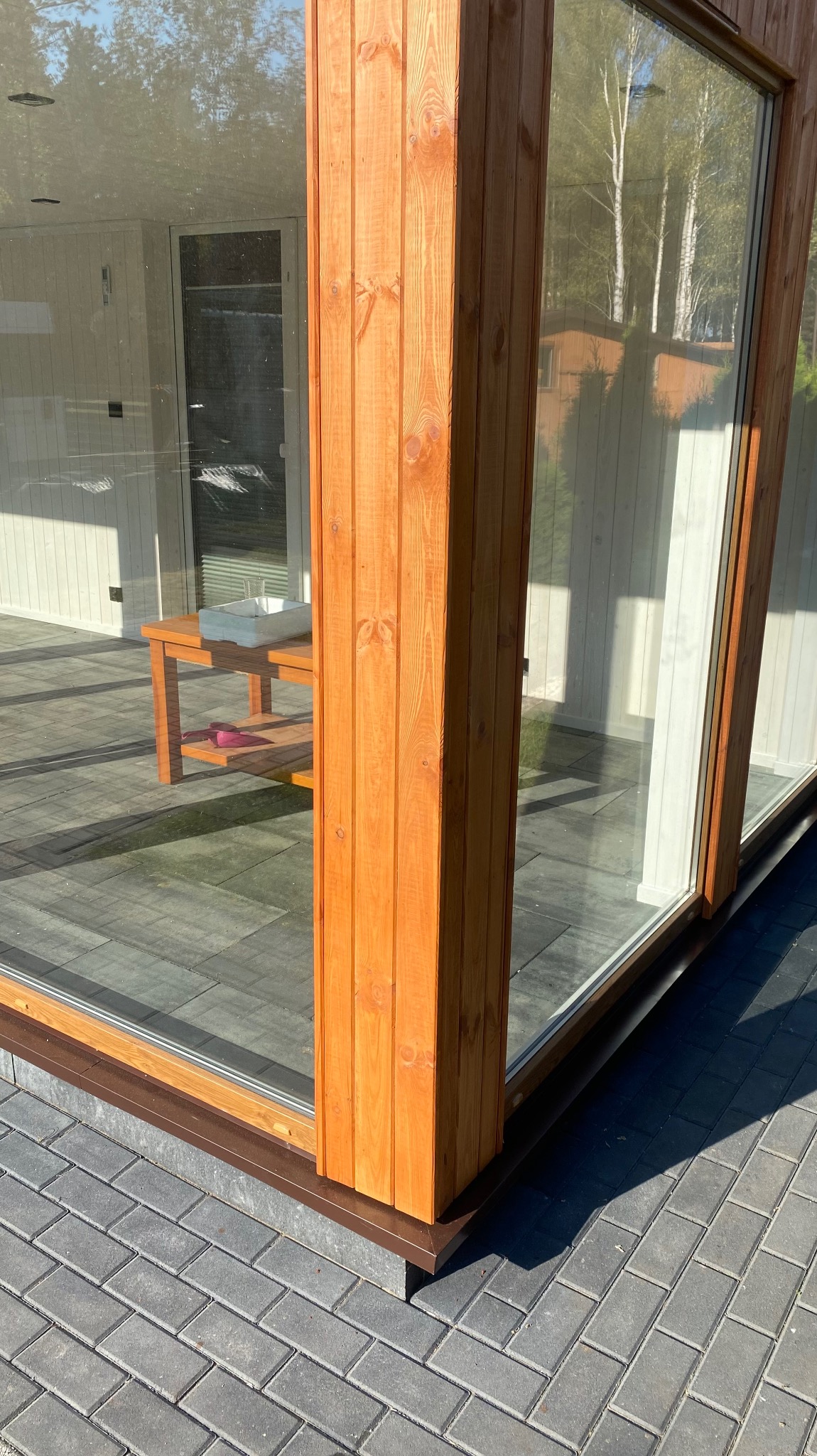 Fragment przeszklonej ściany domu z drewnianą kolumną, widoczny taras z szarej kostki brukowej i odbicie drzew w szybach.
