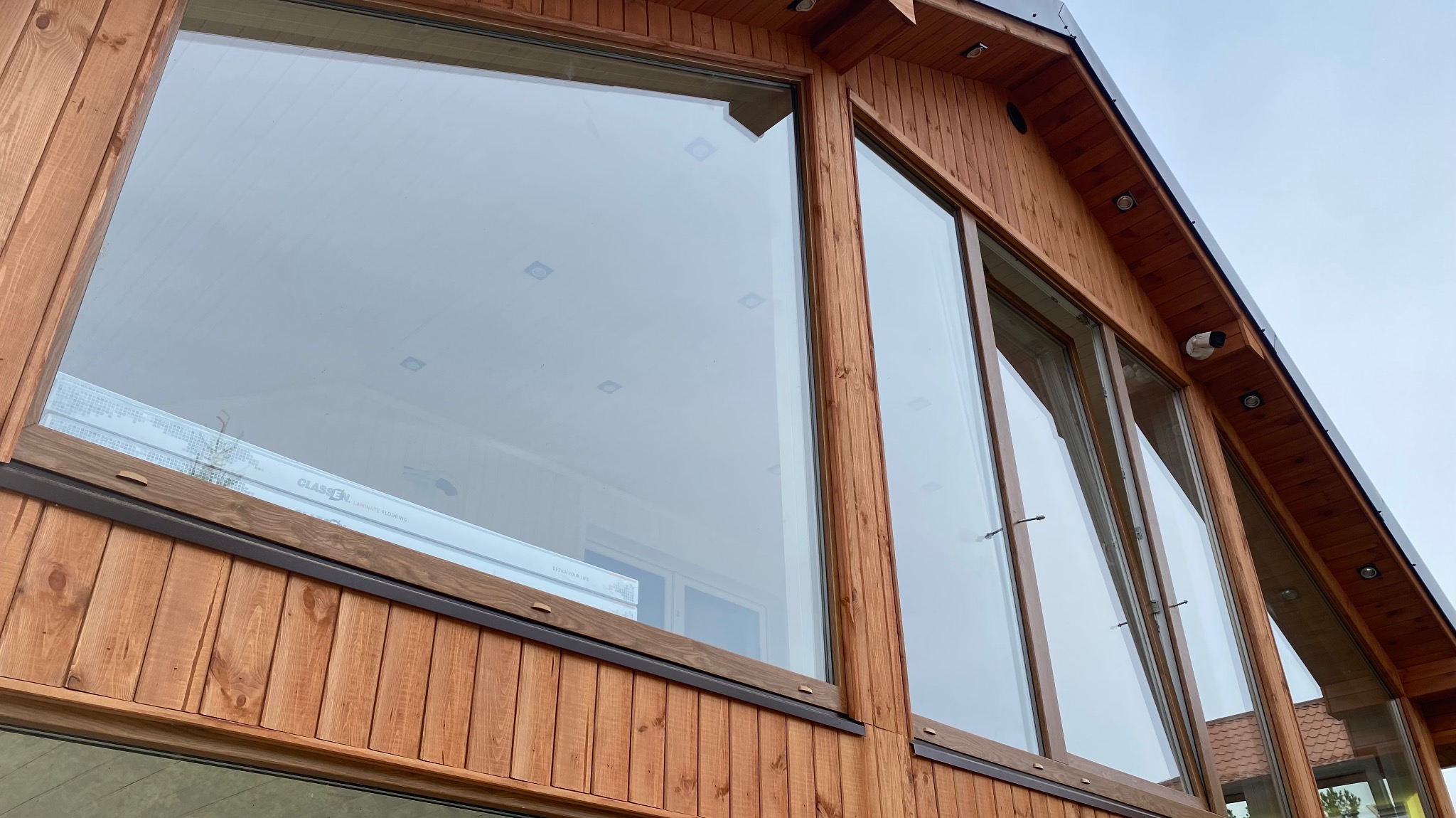 Fragment ściany zewnętrznej domu szkieletowego z dużymi oknami, wykończonego drewnianą boazerią w ciepłym odcieniu brązu, widoczny fragment dachu i oświetlenie zewnętrzne.