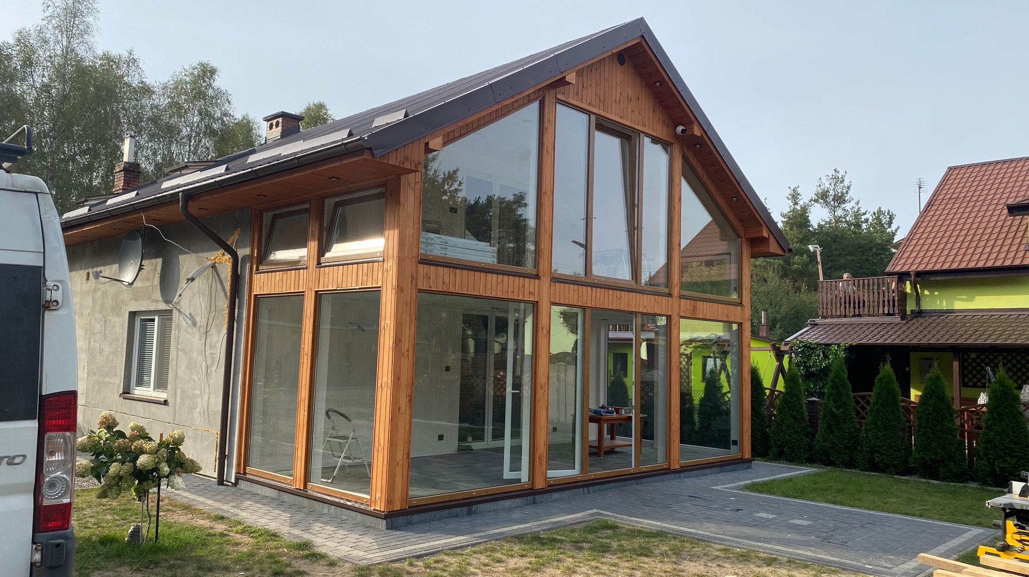 Nowoczesna dobudówka domu z dużymi przeszkleniami i drewnianą konstrukcją, widoczna z zewnątrz, z fragmentem zaparkowanego samochodu dostawczego po lewej stronie i sąsiednim domem w tle.