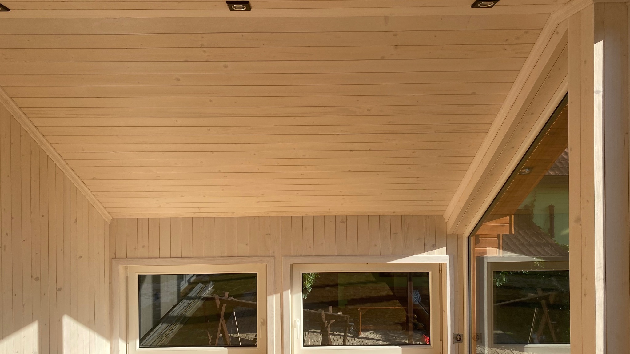Wnętrze jasnej altany z drewnianą boazerią, widoczne okna z odbiciem otoczenia i punktowe oświetlenie sufitowe.
