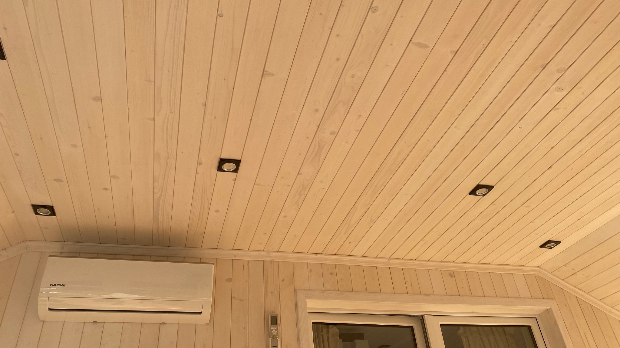 Wnętrze wykończone jasnym drewnem: boazeria na ścianach i suficie, wpuszczone czarne reflektory sufitowe, klimatyzator na ścianie, białe okno.