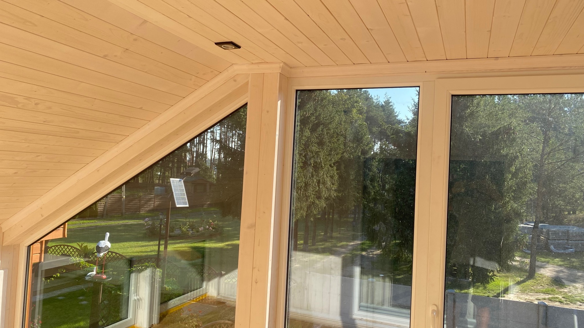 Wnętrze domu z drewnianą boazerią, widok na ogród przez okna z białymi ramami, na zewnątrz dekoracyjny bocian i panel słoneczny.