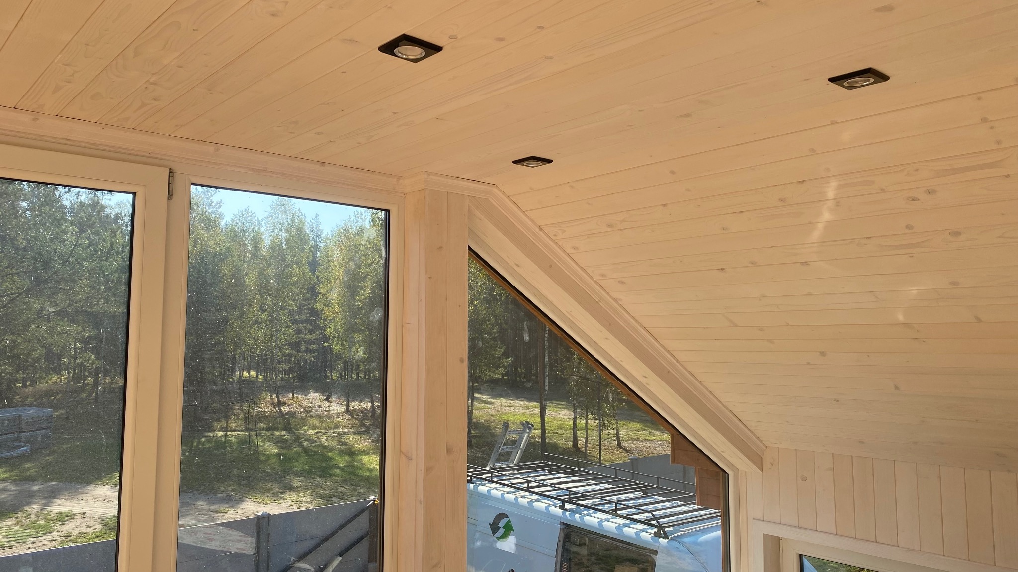 Wnętrze domu w trakcie budowy, widok na drewniany sufit z wbudowanymi reflektorami, duże okno z widokiem na las i samochód z drabinką na dachu.