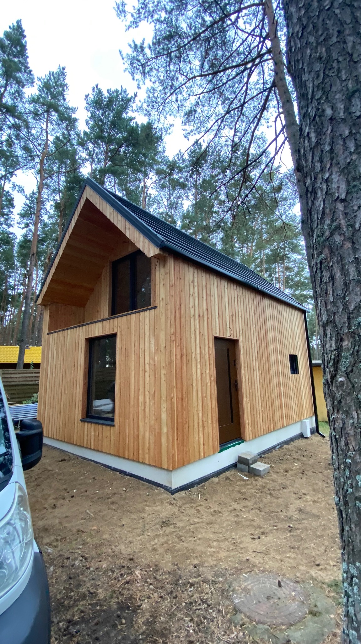 Nowoczesny dom z drewnianą elewacją w otoczeniu drzew iglastych, widoczny fragment białego busa z przodu, surowy teren wokół budynku.