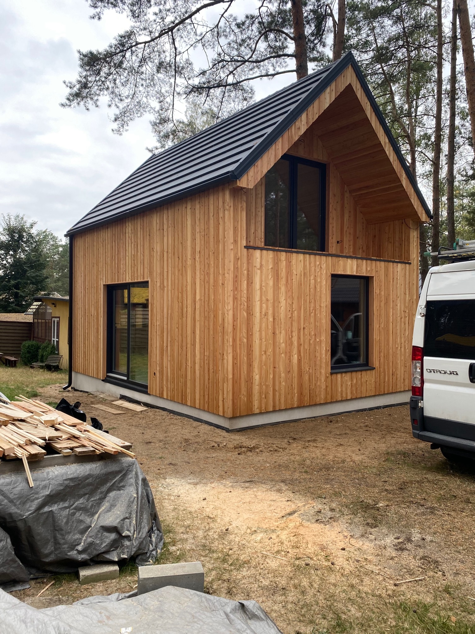 Nowoczesny dom z drewnianą elewacją, czarną dachówką i oknami, widok z zewnątrz na tle lasu, z materiałami budowlanymi i samochodem dostawczym w pobliżu.
