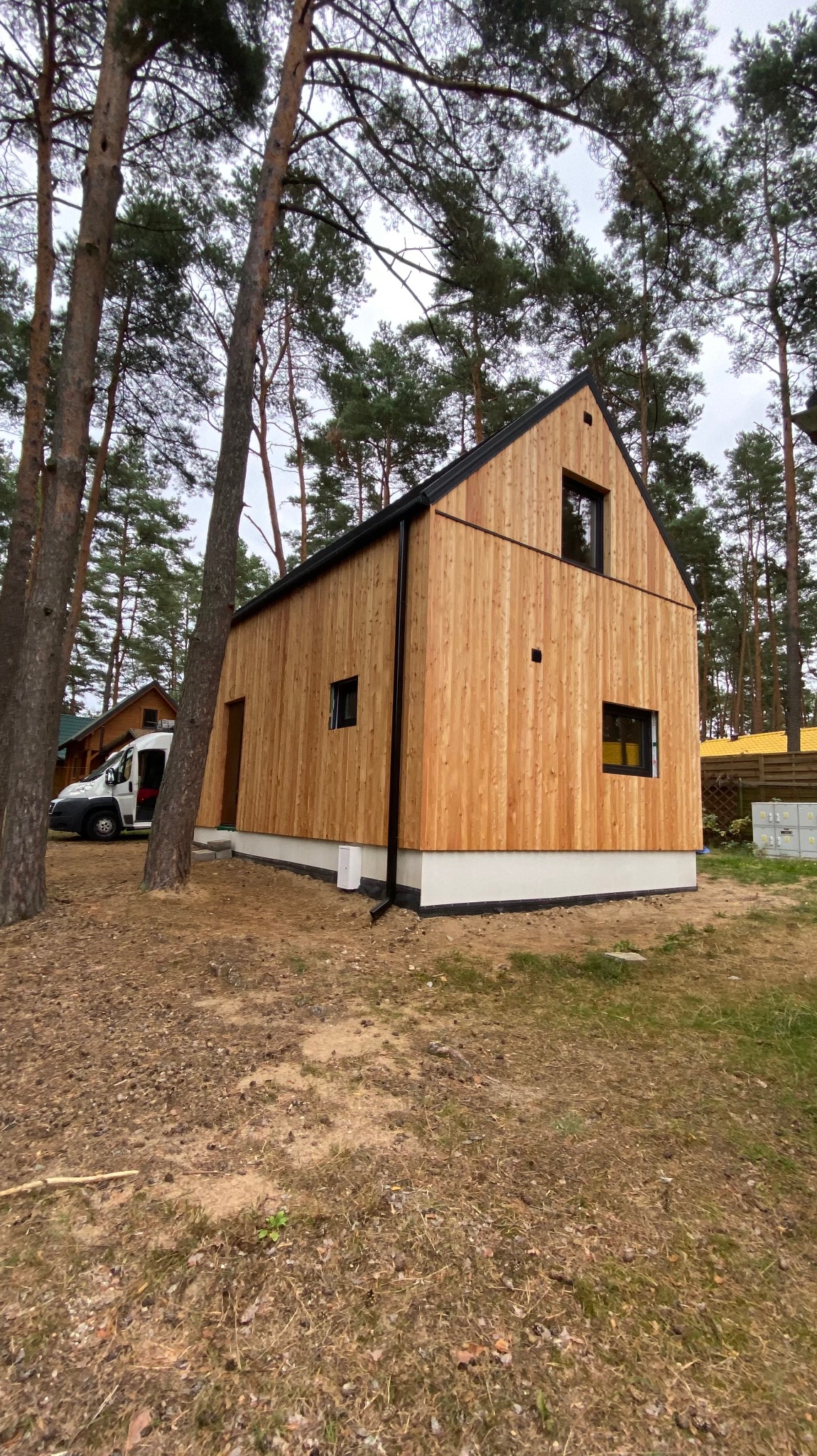 Nowoczesny dom z jasnego drewna w stylu skandynawskim, z czarnymi oknami i dachem, otoczony sosnowym lasem.