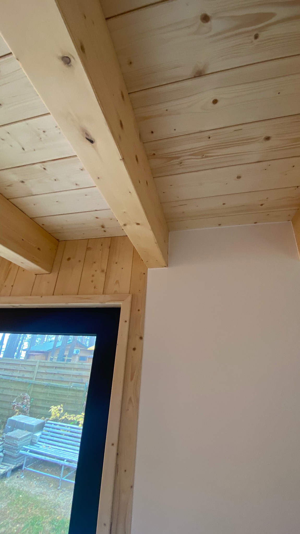 Wnętrze w trakcie budowy, widoczne drewniane belki stropowe, boazeria ścienna i okno z widokiem na ogród z ławką.