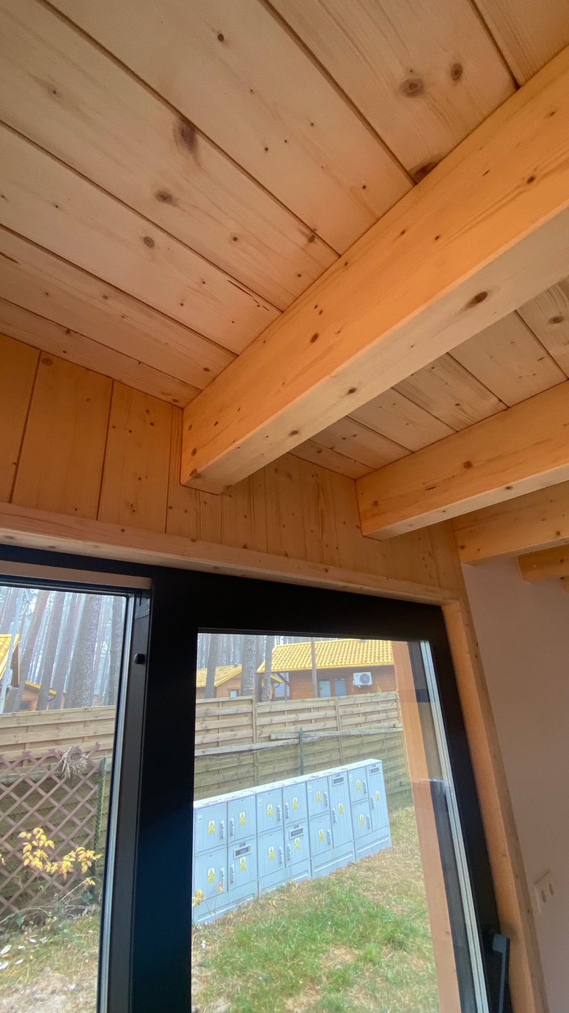 Wnętrze domu szkieletowego w trakcie budowy: widok na drewniany strop z belek, z oknem na zewnątrz gdzie widać skrzynki elektryczne i dom z żółtym dachem.