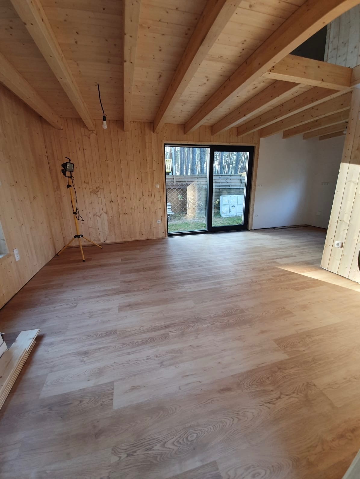 Wnętrze surowego domu w konstrukcji szkieletowej z drewnianymi ścianami i stropem, widoczne okno tarasowe i lampa budowlana na statywie.