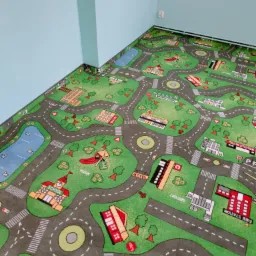 Pokój dziecięcy z wykładziną dywanową imitującą miasto z ulicami, budynkami (szkoła, szpital, remiza strażacka) i parkiem, widok z góry na narożnik pokoju z błękitnymi ścianami.