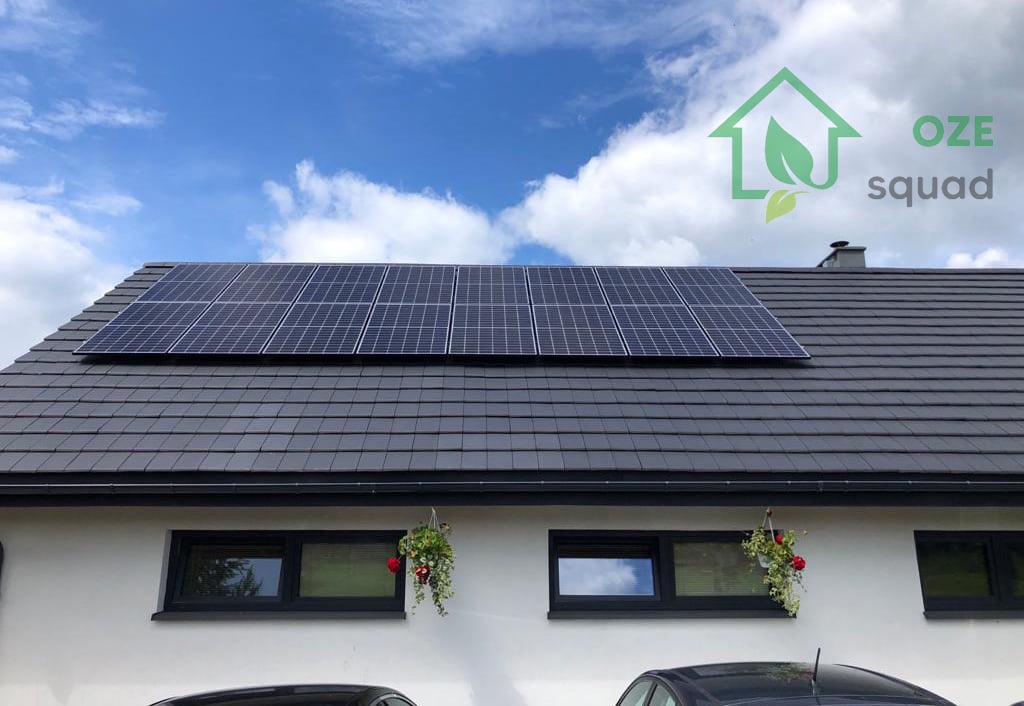 Panele słoneczne na ciemnoszarym dachu domu z białymi ścianami, pod błękitnym niebem z chmurami, z logo OZE Squad w prawym górnym rogu.