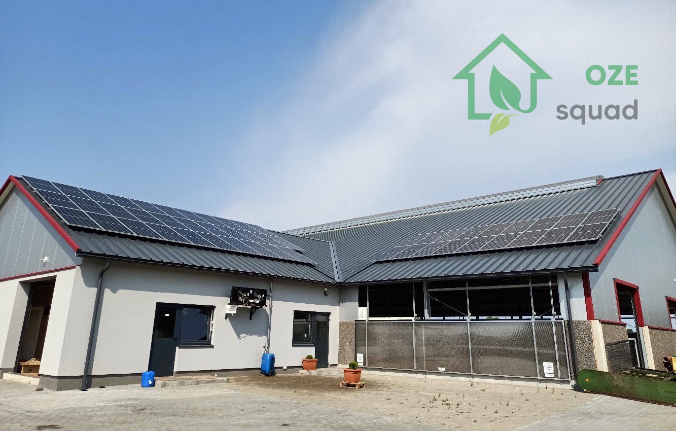 Duży budynek gospodarczy z zamontowanymi panelami słonecznymi na dachu, widoczne również dwa niebieskie pojemniki na zewnątrz budynku oraz logo firmy 'OZE squad' w prawym górnym rogu.