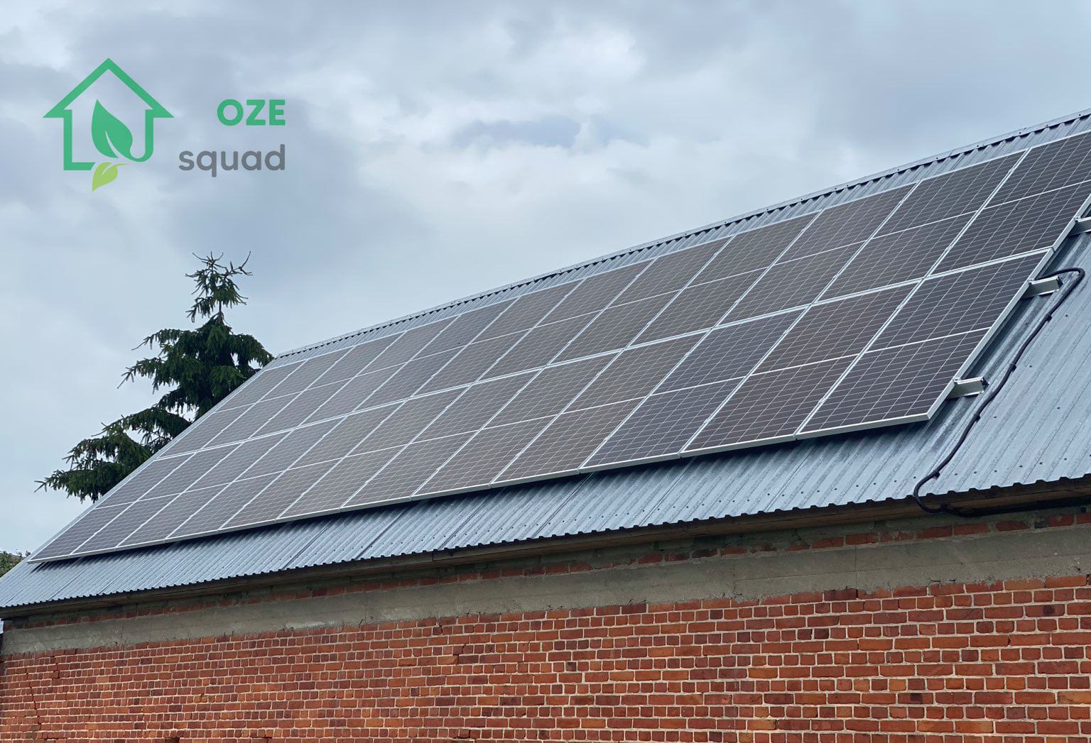Instalacja fotowoltaiczna na metalowym dachu budynku z czerwonej cegły, widoczny fragment logo firmy OZE Squad w lewym górnym rogu.