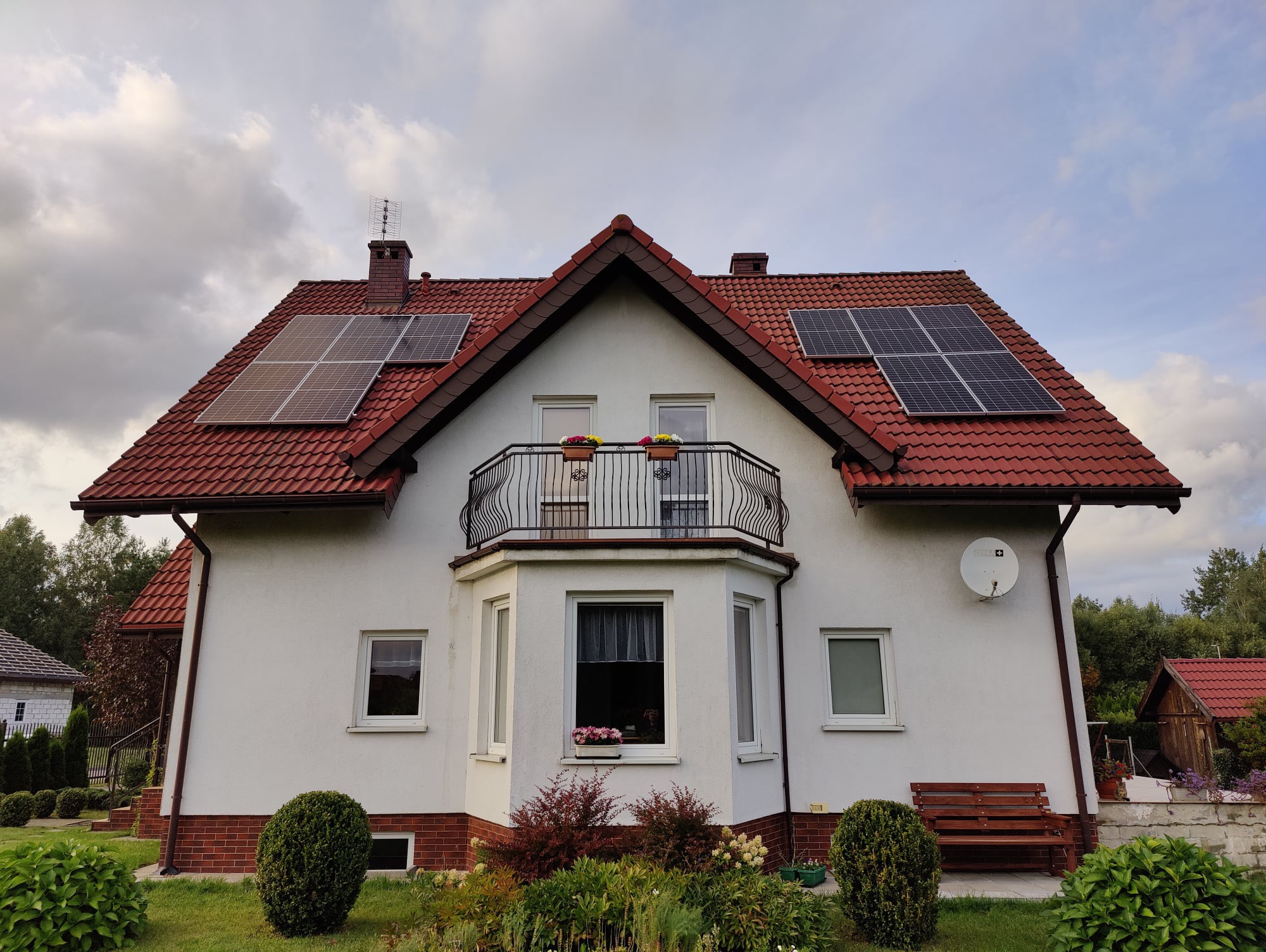 Dwukondygnacyjny dom z czerwoną dachówką, na której zamontowano panele fotowoltaiczne; balkon z kwiatami i zadbany ogród na pierwszym planie.