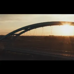 Łukowy most kolejowy o zmierzchu, z promieniami słońca przebijającymi przez konstrukcję i widokiem na miasto w tle.