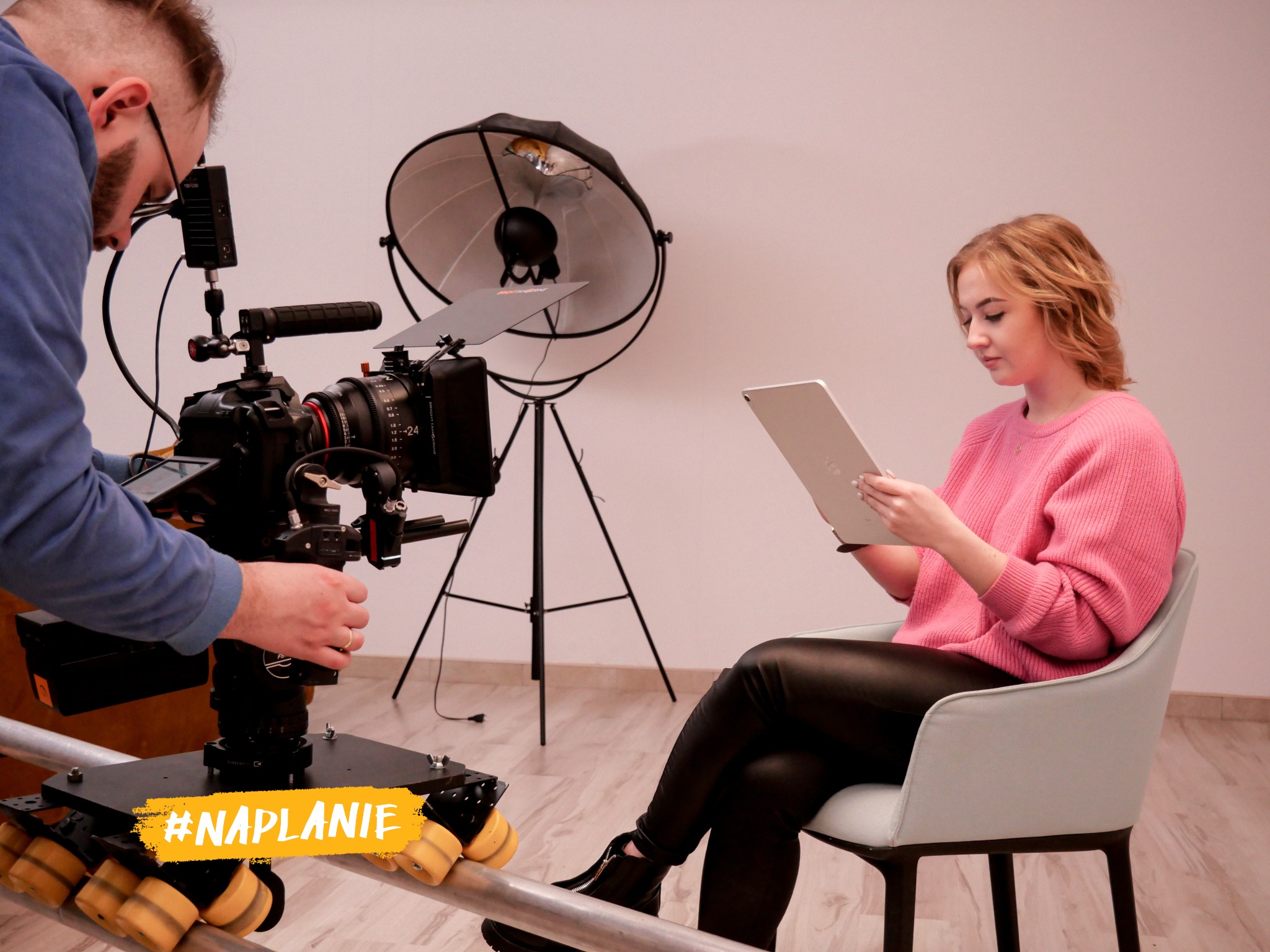 Kobieta w różowym swetrze siedzi na krześle i patrzy na tablet podczas sesji filmowej z profesjonalnym sprzętem oświetleniowym i kamerą na szynach.