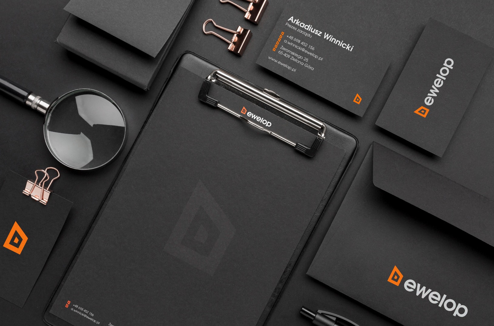 Czarny branding firmy Dewelop z wizytówkami, teczką, kopertami i lupą na czarnym tle, prezentacja identyfikacji wizualnej.