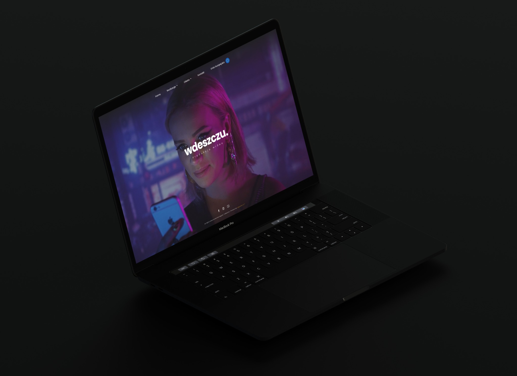 Ekran laptopa MacBook Pro wyświetla stronę internetową z wizerunkiem kobiety i tekstem 'wdeszczu. produkcja wideo' w neonowym oświetleniu.