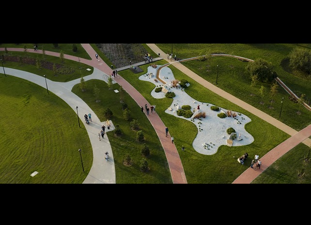Widok z góry na park z alejkami, trawnikami i elementami małej architektury, w tym obszar wysypany białym żwirem z nasadzeniami, otoczony zielenią, z osobami spacerującymi i odpoczywającymi.