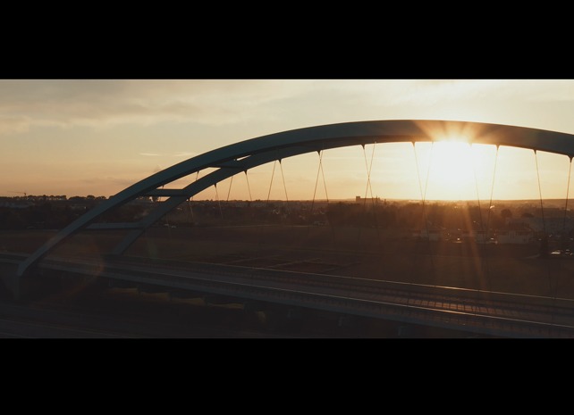 Łukowy most kolejowy o zmierzchu, z promieniami słońca przebijającymi przez konstrukcję i widokiem na miasto w tle.