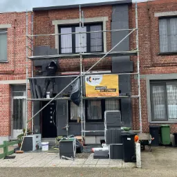 Pracownik na rusztowaniu montuje szare panele izolacyjne na ceglanej elewacji domu jednorodzinnego, widoczny baner reklamowy firmy oraz materiały budowlane na ziemi.