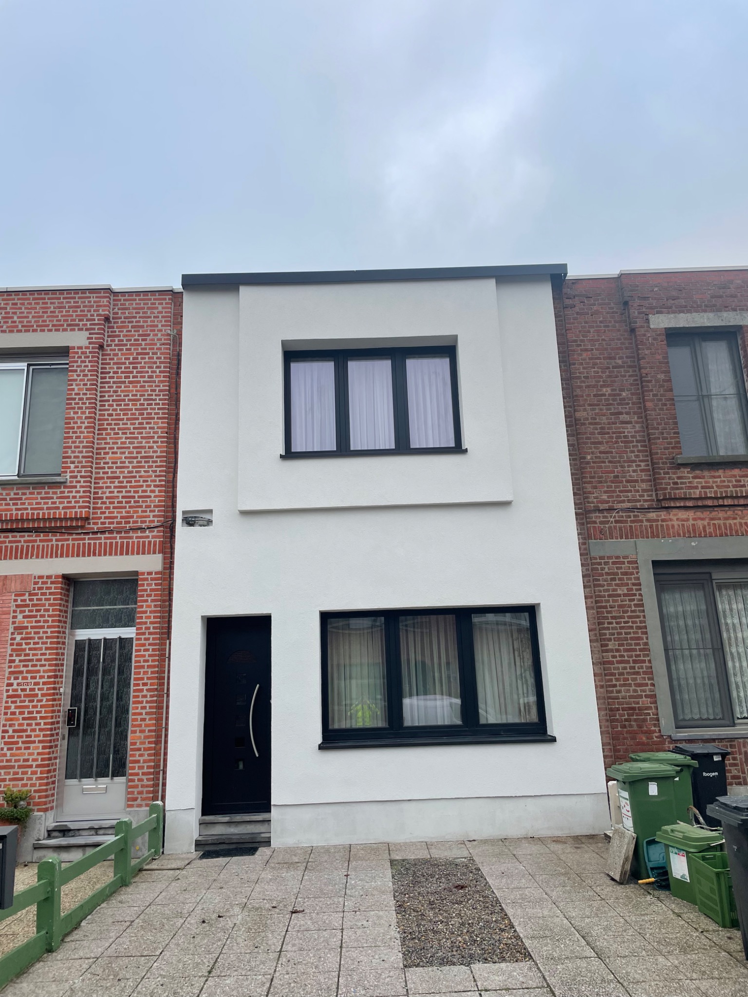 Dwukondygnacyjny dom szeregowy z białą fasadą i czarnymi ramami okiennymi, sąsiadujący z budynkami z czerwonej cegły, widok z poziomu chodnika.