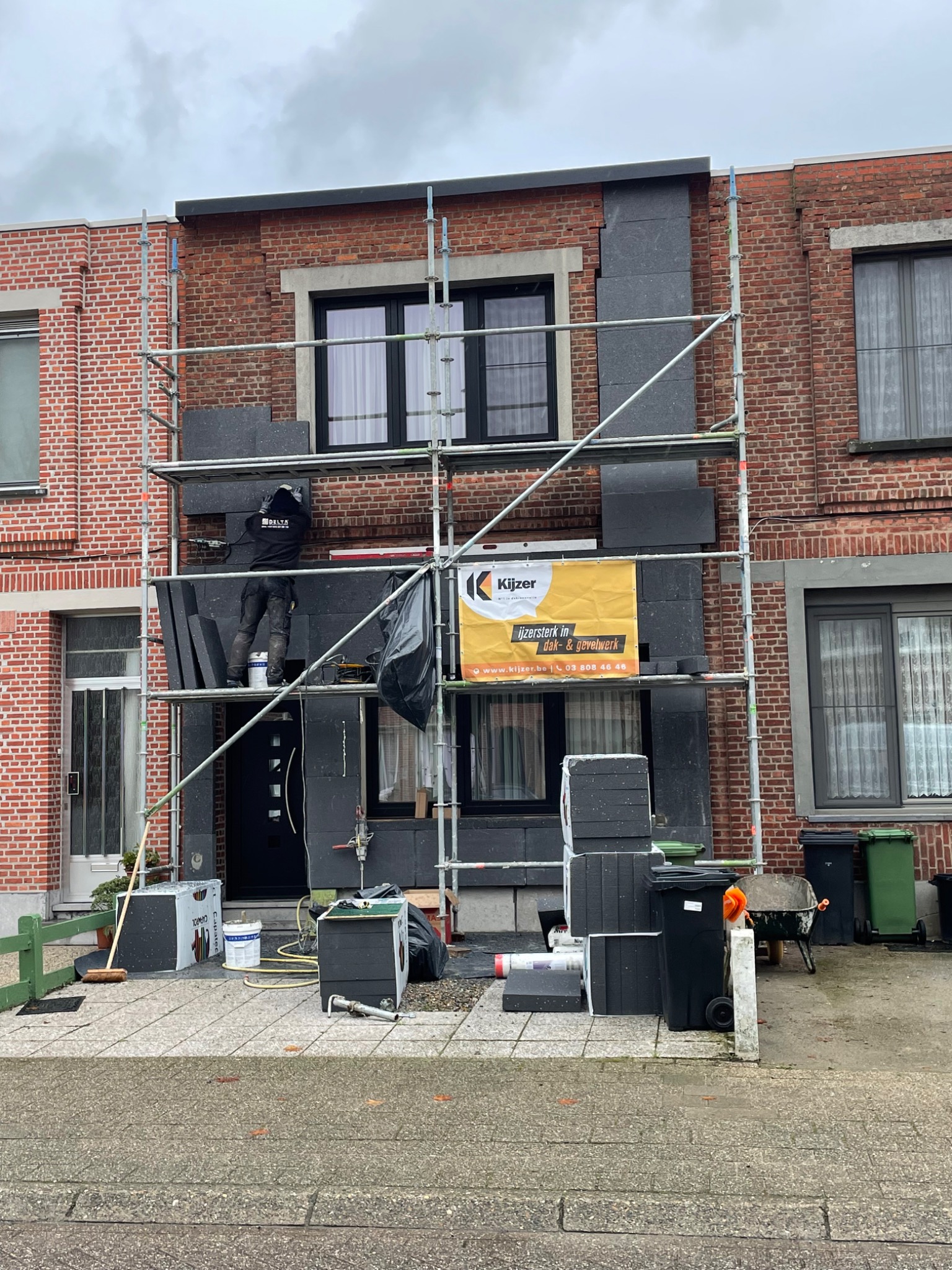 Pracownik na rusztowaniu montuje szare panele izolacyjne na ceglanej elewacji domu jednorodzinnego, widoczny baner reklamowy firmy oraz materiały budowlane na ziemi.