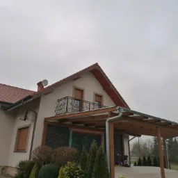 Dwupiętrowy dom jednorodzinny z beżową elewacją, drewnianą pergolą i balkonem z ozdobną, kutą balustradą, widoczny z perspektywy ogrodu w pochmurny dzień.