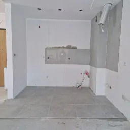 Wnętrze w trakcie prac: szare płytki podłogowe, fragment ściany z położonymi płytkami, widoczne ślady po kleju, otwory w suficie, puszka elektryczna oraz wiadro z resztkami zaprawy.