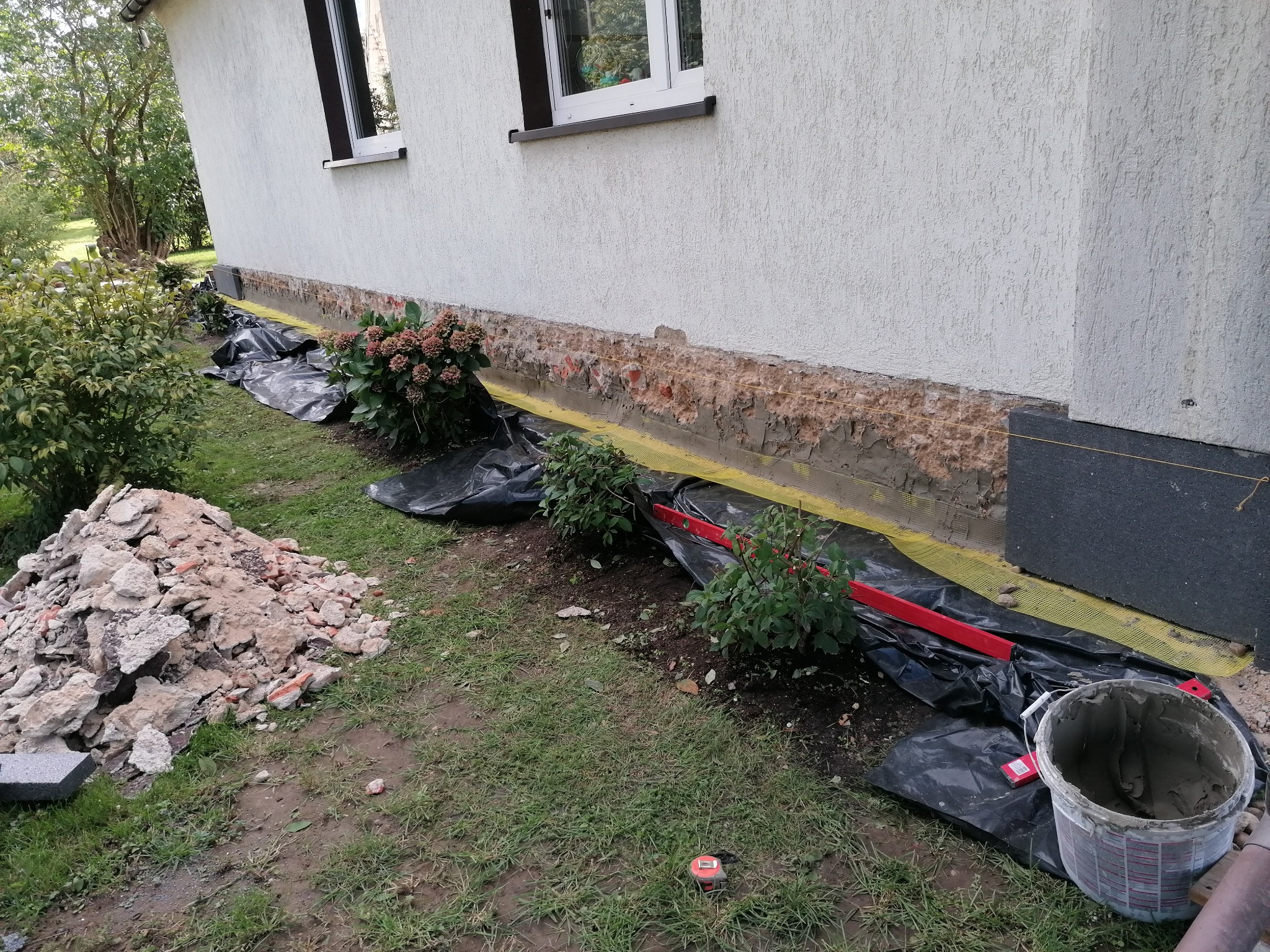 Przygotowanie do ocieplenia fundamentów budynku: odkryte fundamenty, zabezpieczone folią, widoczny stos gruzu, poziomica i wiadro z zaprawą. Etap prac przed nałożeniem izolacji.