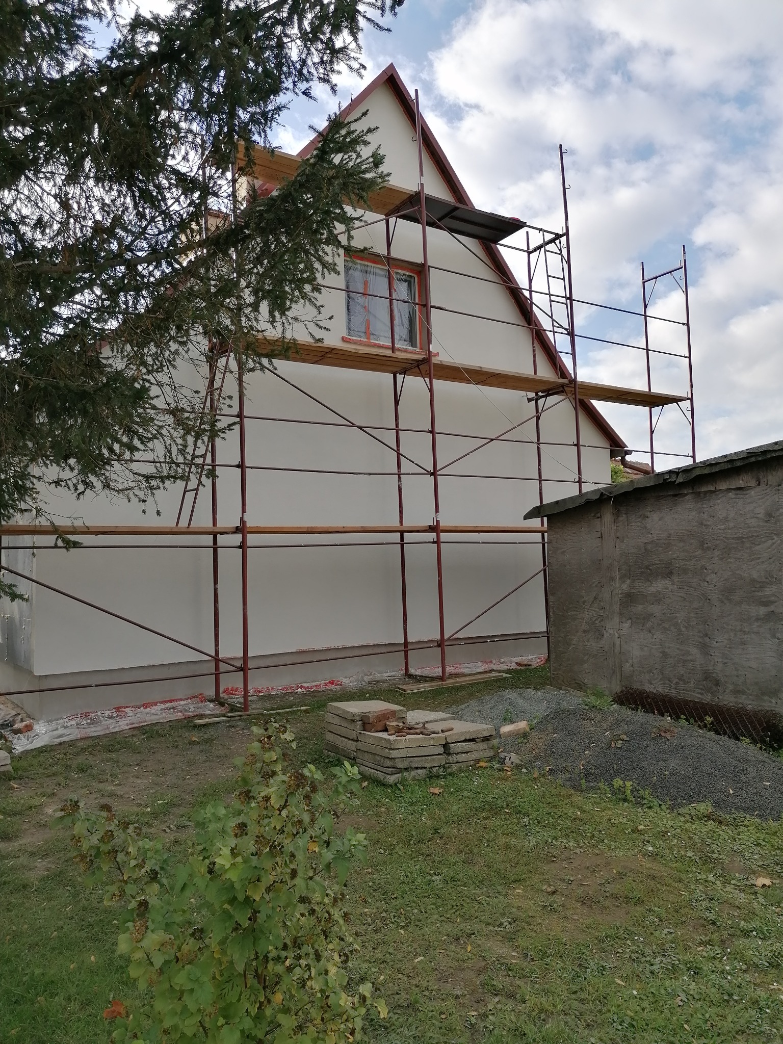 Dom jednorodzinny w trakcie prac elewacyjnych, widoczne rusztowanie, zabezpieczone okno i świeżo otynkowana ściana boczna, na trawniku sterta gruzu i materiałów budowlanych.